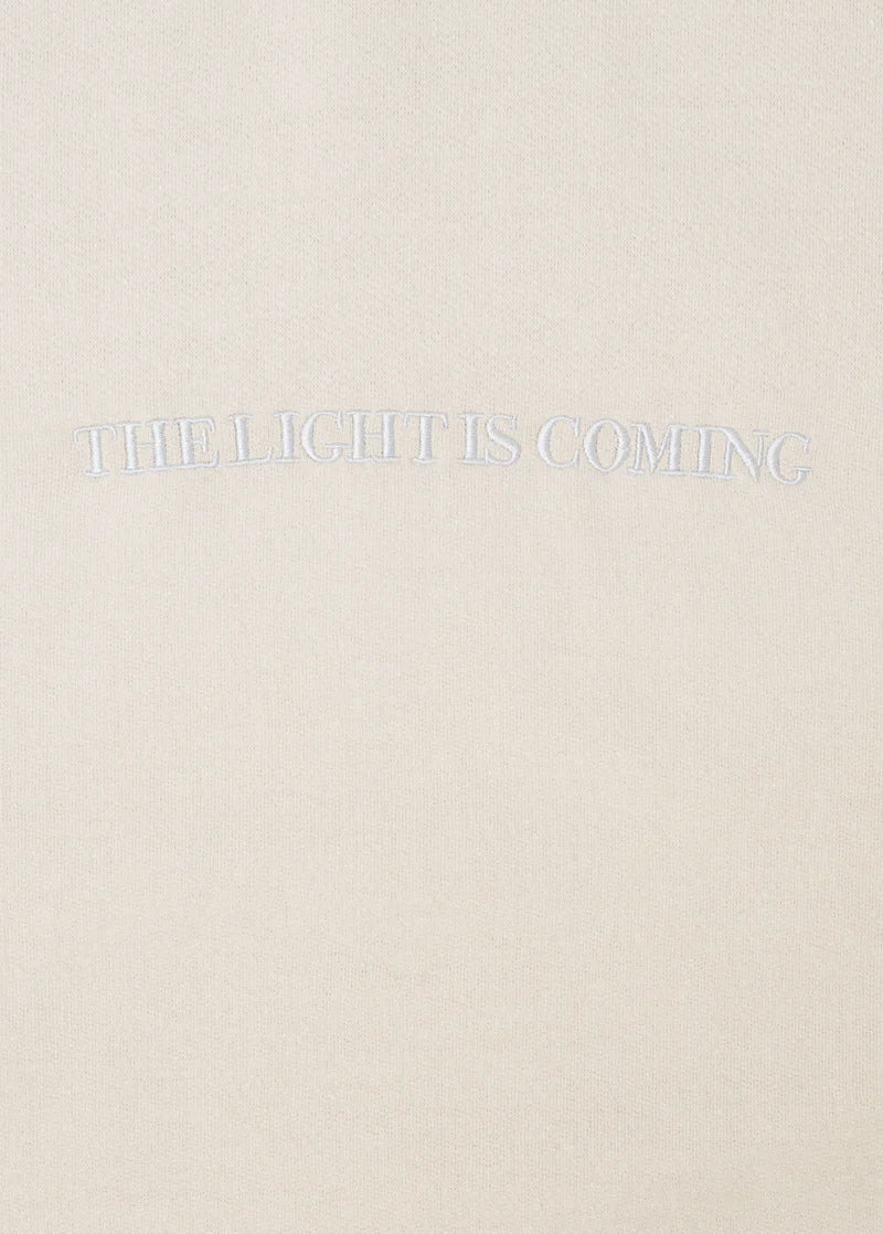 Close-up of embroidered The Light Is Coming text on cream Ariana Grande crewneck sweatshirt with tonal stitching
